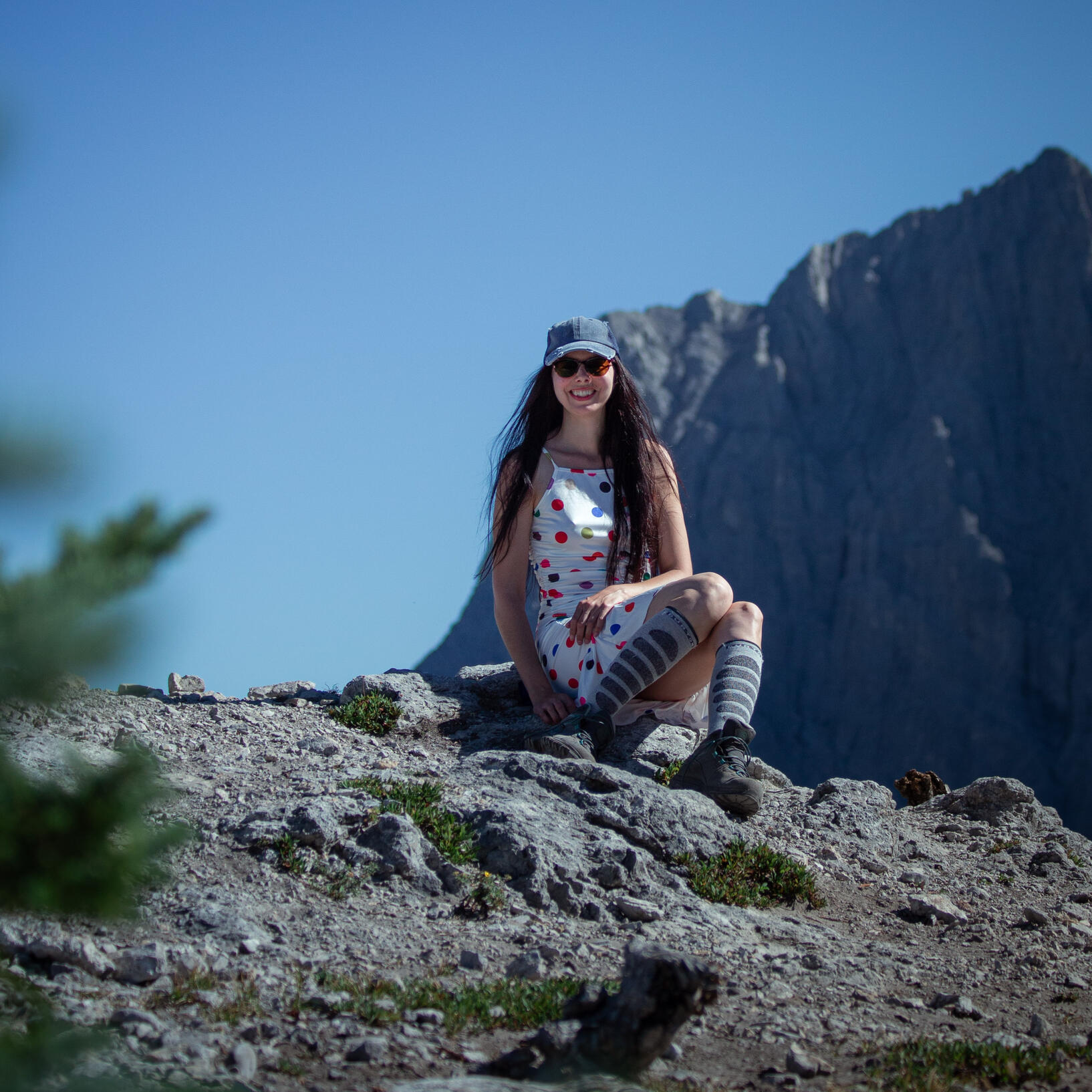 A happy brunette in a polkadot dress, a denim ball cap, and hiking boots sitting on a rock in the alpine.